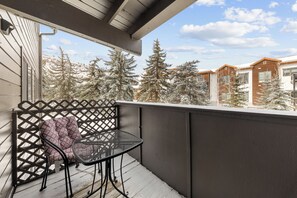 A balcony with a metal table and a cushioned chair, overlooking snow-covered trees and neighboring buildings. - Near the slopes in Avon! (Avon)