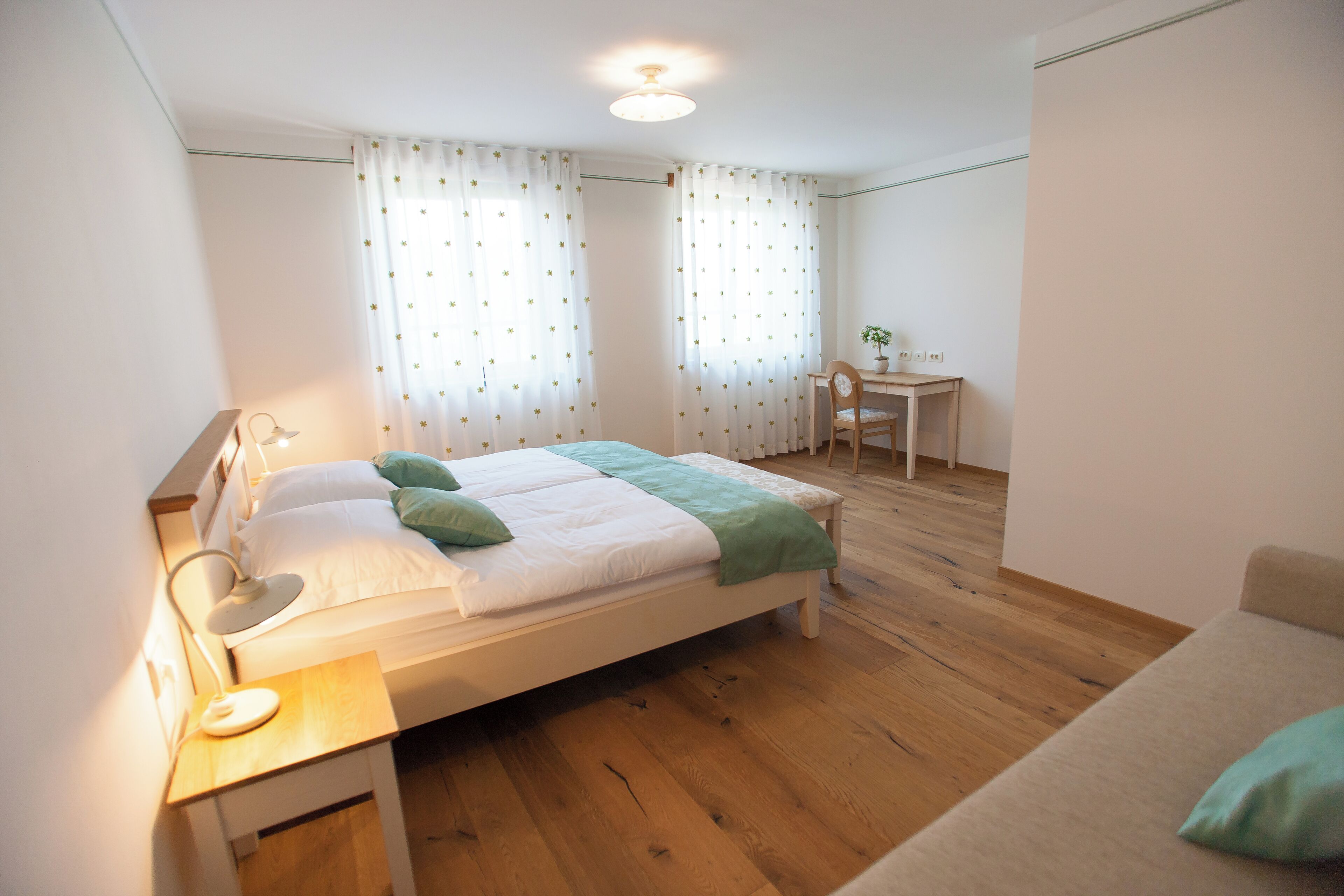 A bedroom with a bed, bedside table, lamp, curtains, a desk, chair, and a small potted plant.