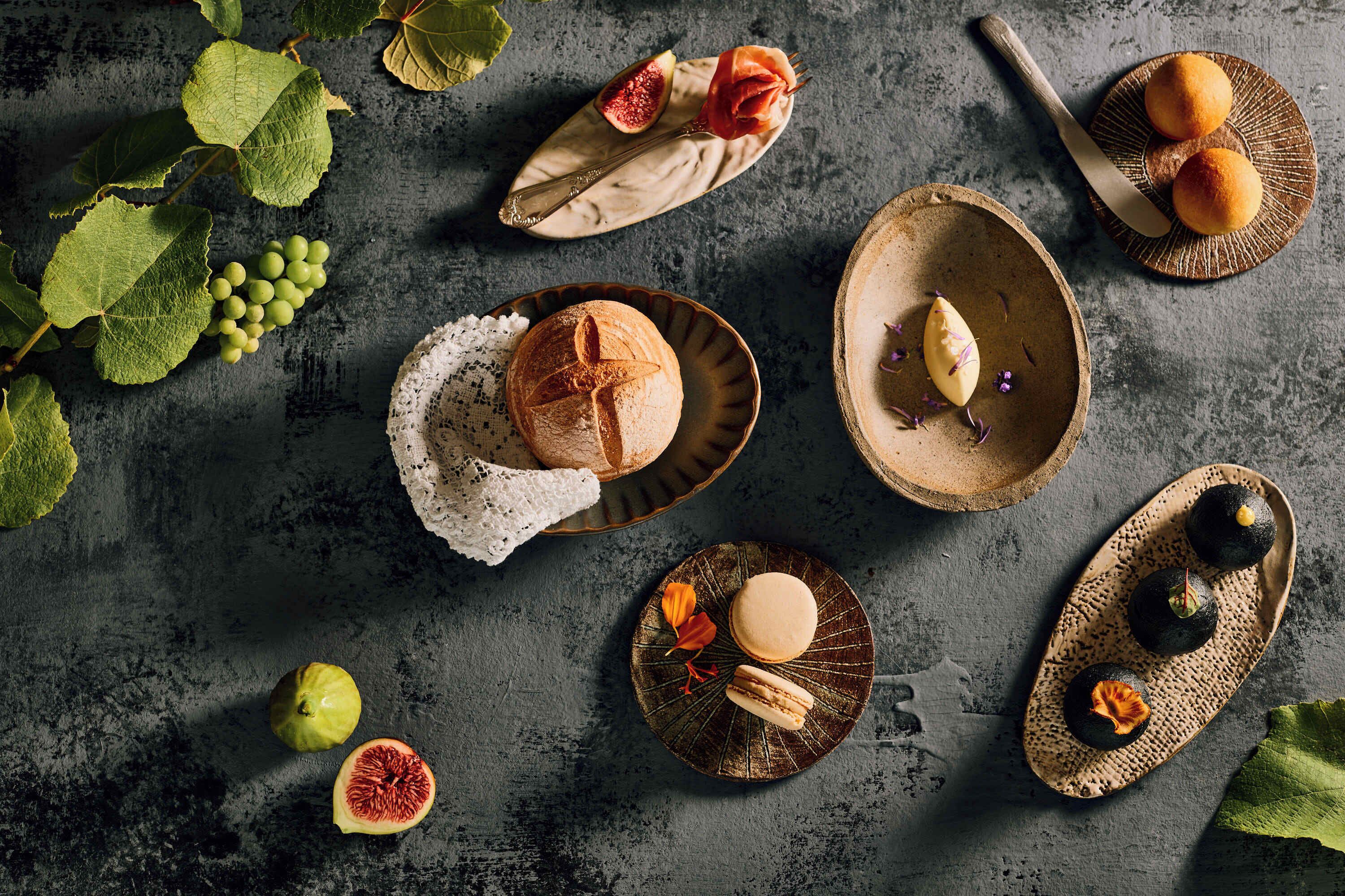 Uma mesa rústica com pão, figos e macarons sobre uma superfície texturizada.