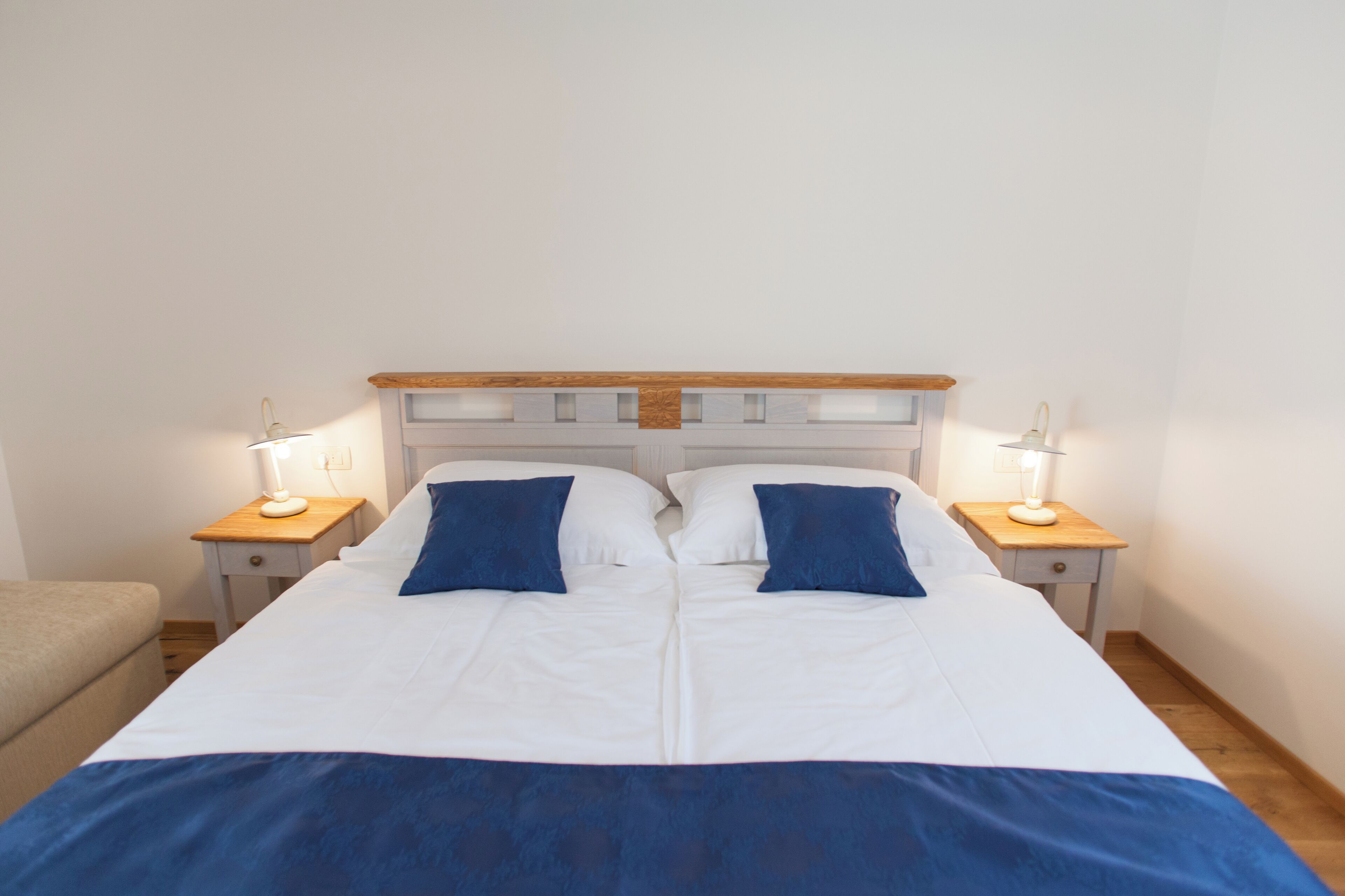Uma cama de casal com roupa de cama branca e azul, duas luminárias de cabeceira e uma cabeceira de madeira.