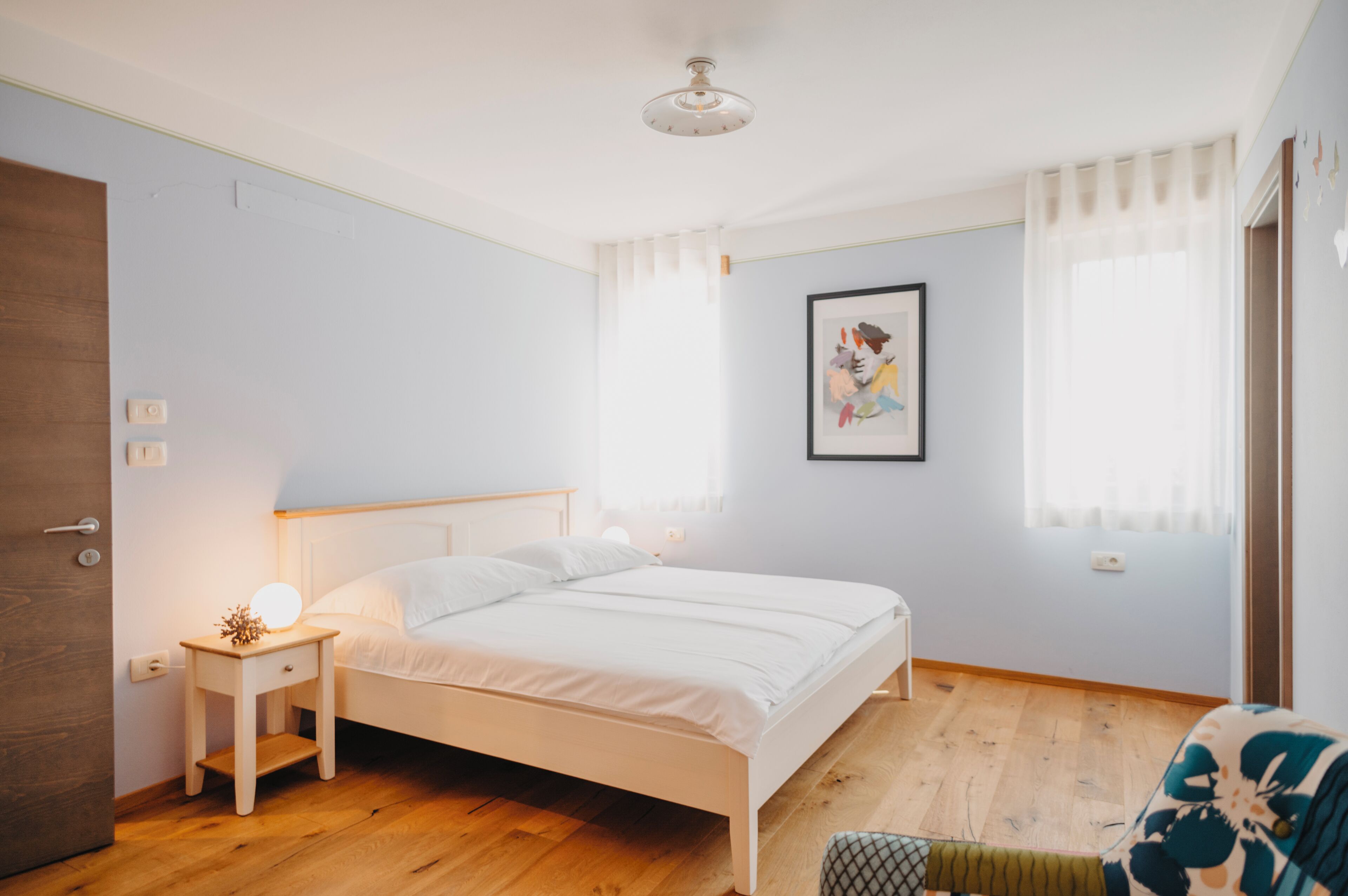 A bedroom with a wooden floor, a white bed, a bedside table with a lamp, a framed picture on the wall, and a window with sheer curtains.