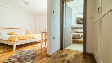 Ein Schlafzimmer mit Bett, Nachttisch, Stuhl, einem farbenfrohen Teppich und Blick ins Bad mit Dusche und Waschbecken.