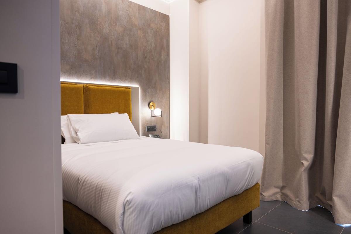 Une chambre d’hôtel comprenant un lit, une tête de lit, une table de chevet, une lampe fixée au mur et un rideau.