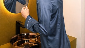 Una persona con camisa azul maquillándose frente a un lavamanos con un espejo ovalado grande y un recipiente metálico.