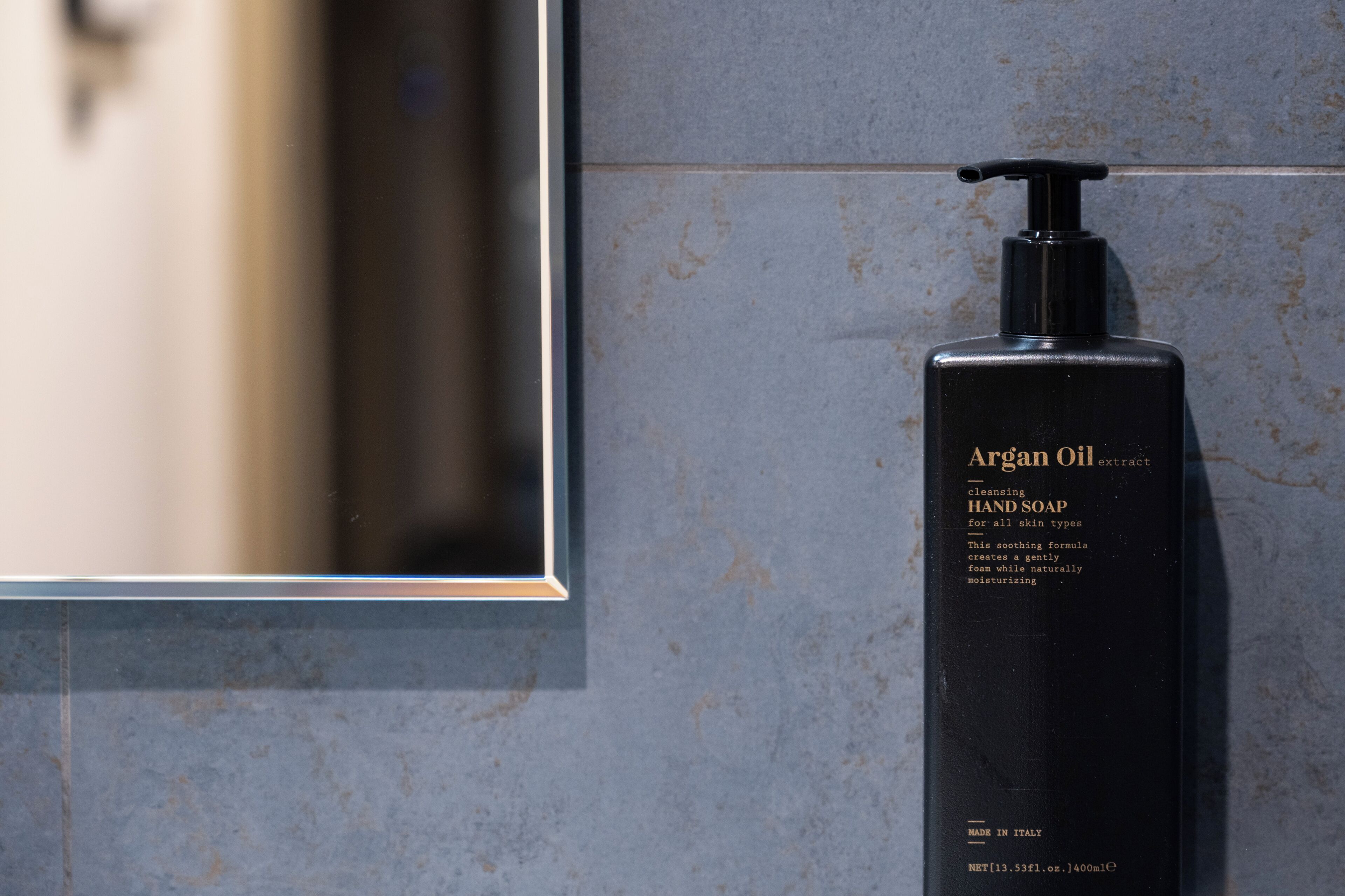 En sort sæbebeholder til håndsæbe med påskriften 'Argan Oil extract' på en flisevæg.