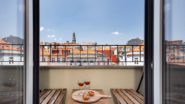 A balcony with a wooden table, two glasses of wine, and a pastry on a cutting board, overlooking a cityscape with buildings and a tower. - Comfy 3BR Duplex in Historical Center (Oporto)