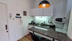 A compact kitchen with white cabinets, a granite countertop, a black chair, and a hanging pot rack. - Studio 325 Teresópolis - Best Location.  WATERFALLS AND GASTRONOMY (Teresópolis)