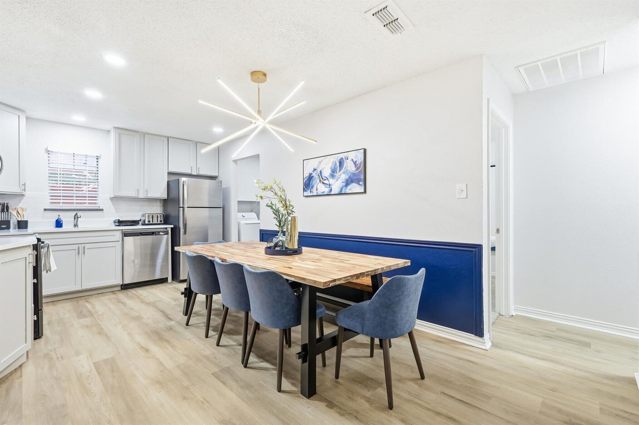 A modern kitchen with white cabinets, a stainless steel refrigerator, and a wooden dining table with blue chairs.