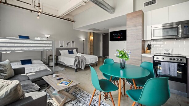 Et moderne hotelværelse med køjeseng, sofa, spisebord med stole, et tekøkken med mikroovn og ovn, samt et fladskærms-tv på væggen.