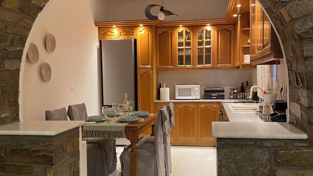 A stone-arched kitchen with wooden cabinets, a dining area with a table and chairs, and a microwave on the counter. - Deppy's House (Naousa)
