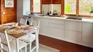 L’intérieur d’un chalet accueillant, avec un plafond en bois, un coin repas doté de chaises blanches, une cuisine avec un réfrigérateur blanc et des fenêtres offrant une vue sur la verdure.
