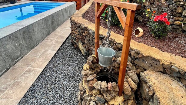 Une structure en bois abritant un puits en pierre et un seau en métal, le tout entouré d’un jardin aux fleurs multicolores et d’une piscine.