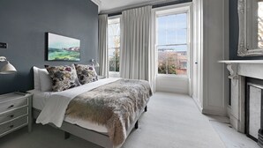 A bedroom with a large bed, a nightstand, a lamp, a painting, and a view of the outside through a window. - Compton Terrace N1 🚇 Highbury and Islignton (London)