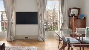 A living room with a large flat-screen TV mounted on the wall, a dining table with chairs, a wooden cabinet with a mirror, and a view of the outside through large windows with curtains. - Compton Terrace N1 🚇 Highbury and Islignton (London)