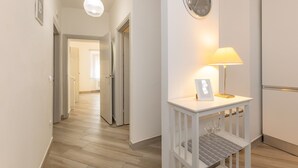Ein Korridor mit einer weißen Tür, einer Wanduhr, einem Tisch mit einer Lampe und einem gerahmten Bild.