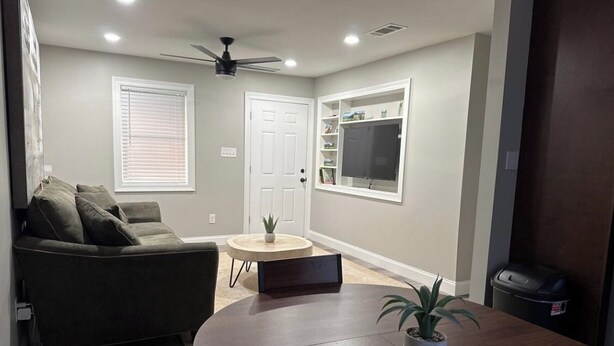 Una sala de estar con un comedor de madera oscura, un sofá, una mesita redonda con una planta, un ventilador de techo y un televisor montado en la pared.