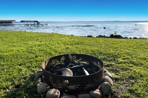 A metal fire pit with rocks, surrounded by grass, overlooking a body of water with a dock and boats. - Alden East Torch Estate (Alden)