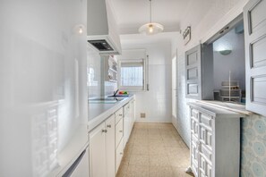 Eine schmale Küche mit weißen Schränken, einer gefliesten Spritzschutzwand und einem Fenster mit Jalousien.