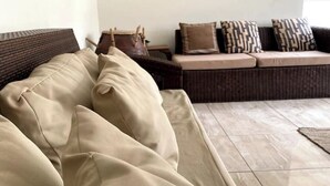 Un sofá beis con varios cojines, una silla de mimbre y un juego de cojines estampados sobre un suelo de baldosas.