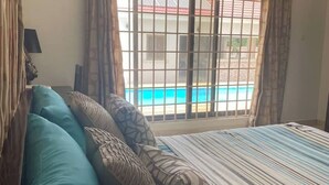 Un dormitorio con cama, almohadas, una ventana y vista a una piscina y un edificio.
