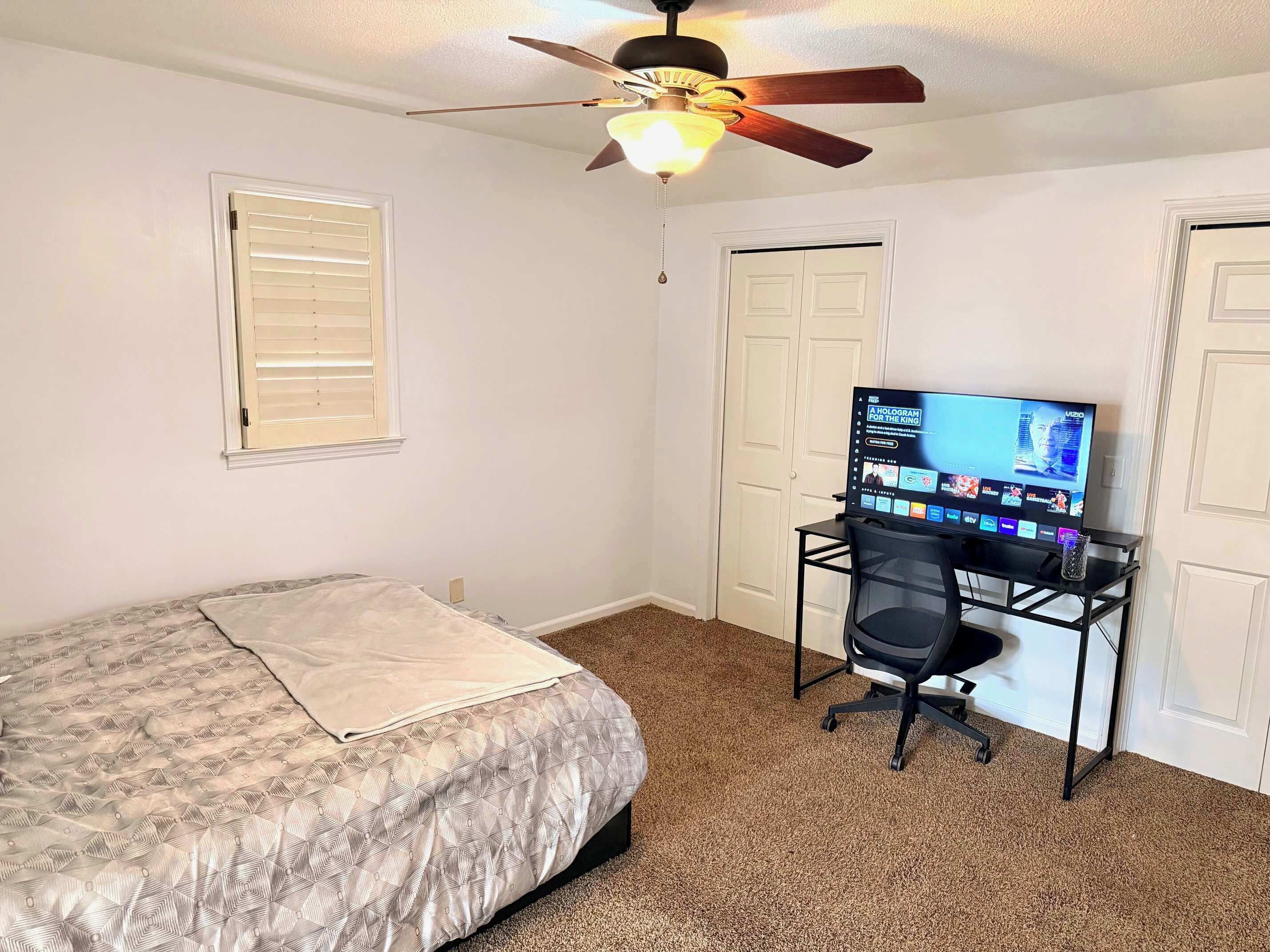 A bedroom with a bed, a desk with a computer, a ceiling fan, and a door.