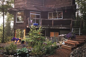 Eine zweistöckige Holzhütte mit Balkon, umgeben von einem Garten mit verschiedenen Pflanzen und einer Holzterrasse.