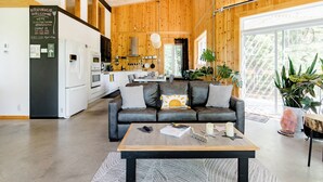 Una cocina moderna con refrigerador, horno y lavamanos, una sala de estar con sofá y mesa de centro, y un ventanal con vista a la vegetación.