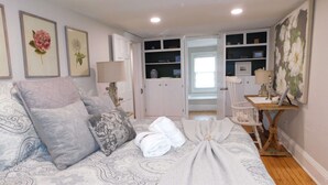 Ein Schlafzimmer mit Bett, Nachttischen, Schreibtisch und Stuhl. An den Wänden hängen Bilder in Rahmen, ein Fenster und eine Tür zu einem weiteren Raum.