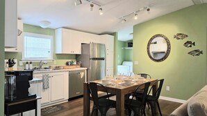 Cocina compacta con armarios blancos, refrigerador de acero inoxidable y comedor con mesa y sillas de madera.