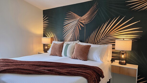 Una cama bien tendida con sábanas blancas y una manta marrón. Hay dos mesitas de noche con lámparas, y un mural grande en la pared con hojas de palmera.