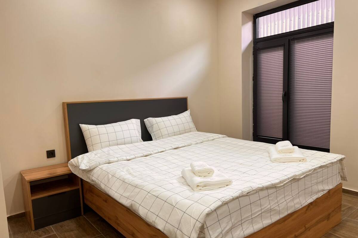 Een net opgemaakt bed met een geruit beddegoed en twee opgevouwen handdoeken, een houten nachtkastje en een raam met jaloezieën.