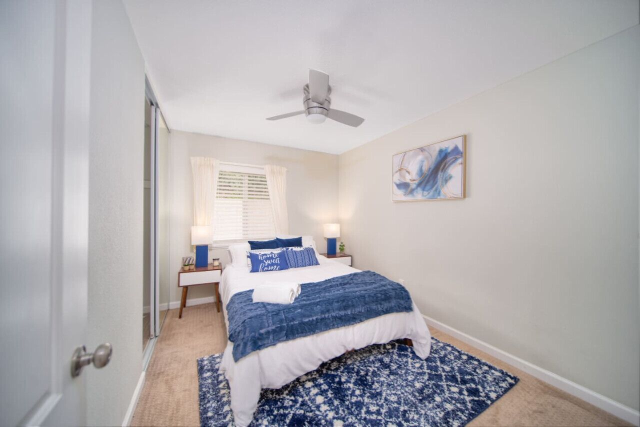 A bedroom with a bed, a ceiling fan, a window with blinds, a nightstand, a lamp, and a painting on the wall.