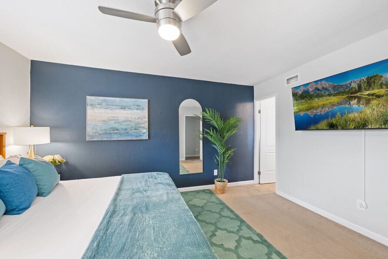 A bedroom with a bed, a ceiling fan, a painting, a mirror, a potted plant, and a view of a lake through a window.