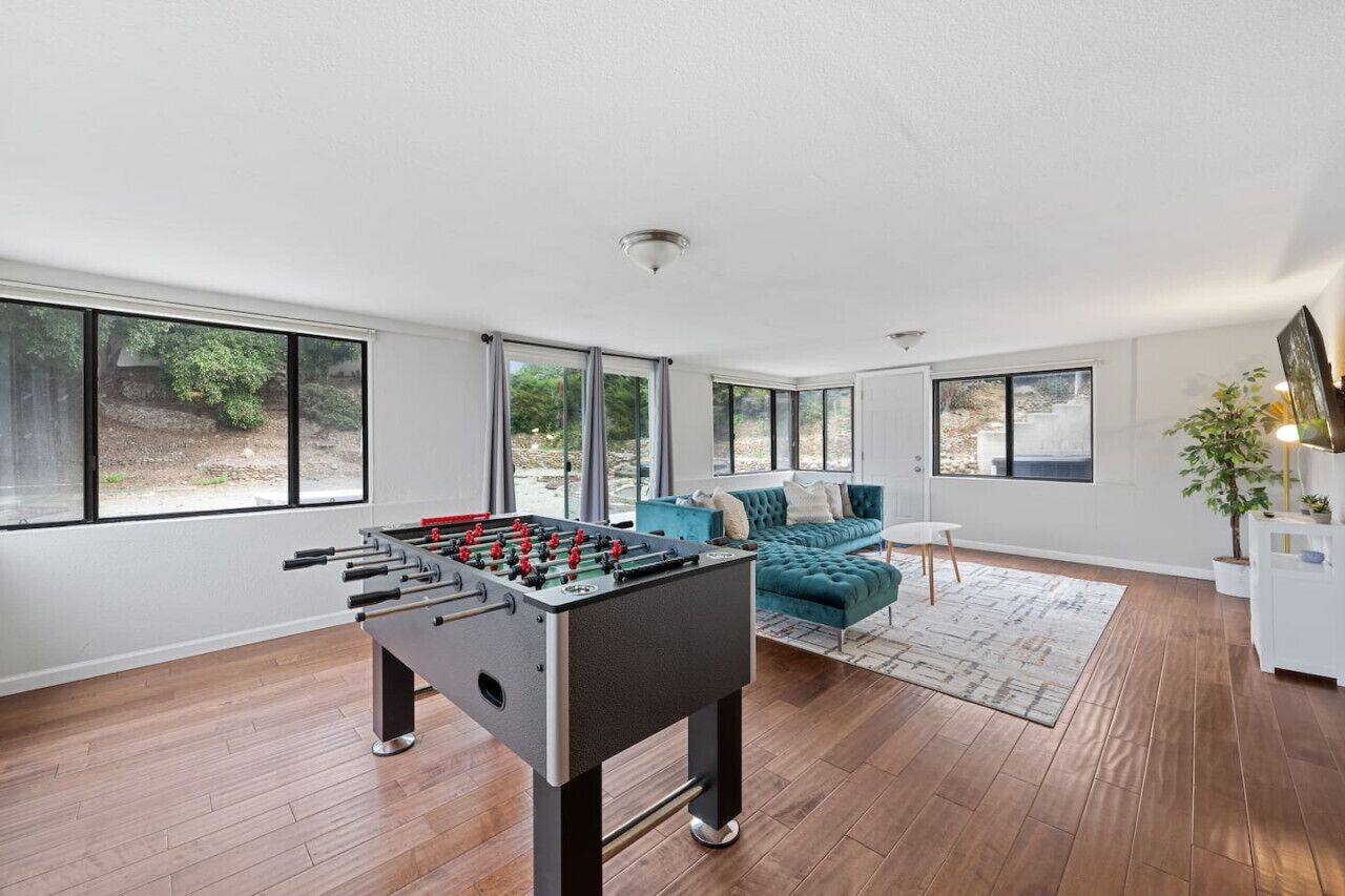 Una sala de estar moderna con mesa de fútbol, un sofá color turquesa y ventanas amplias con vistas al exterior.