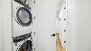 A laundry room with two front-loading washing machines and dryers, white tiled walls, and a wooden floor. - Steps to Transit • Bright Kitchen • Parks & Shops (Calgary)