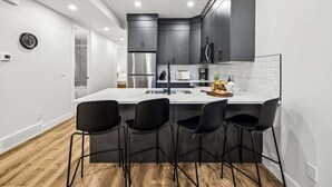 A modern kitchen with dark cabinets, a white countertop, and black bar stools. - Steps to Transit • Bright Kitchen • Parks & Shops (Calgary)