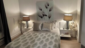 Ein ordentlich eingerichtetes Schlafzimmer mit einem Bett, zwei Nachttischen, einer Wandlampe und einem Blumenbild.