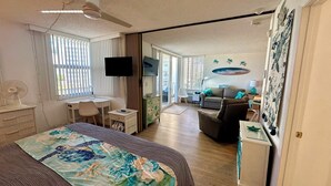 A bedroom with a bed, a desk, a chair, a TV, and a sliding door leading to a living area with a sofa and a coffee table. - Estero Beach & Tennis 205B - Beach View (Fort Myers Beach)