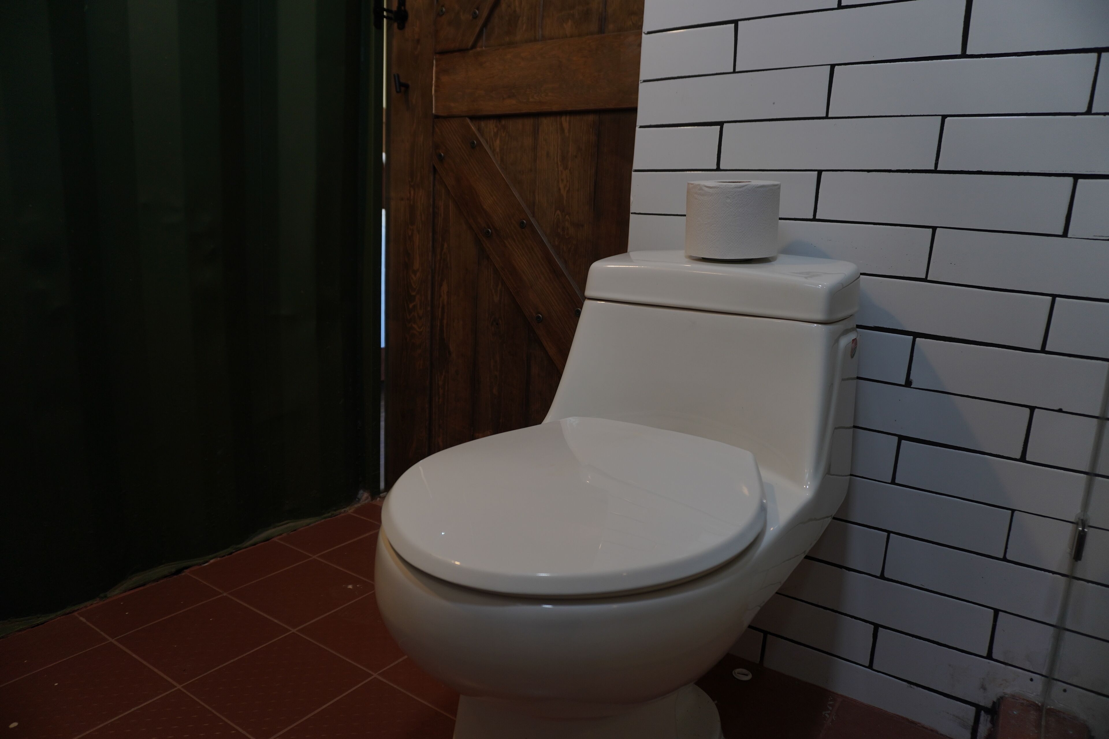 Un inodoro blanco con un rollo de papel higiénico encima, frente a una pared con azulejos blancos y una puerta de madera.