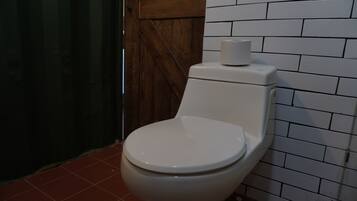 Un inodoro blanco con un rollo de papel higiénico encima, frente a una pared con azulejos blancos y una puerta de madera.