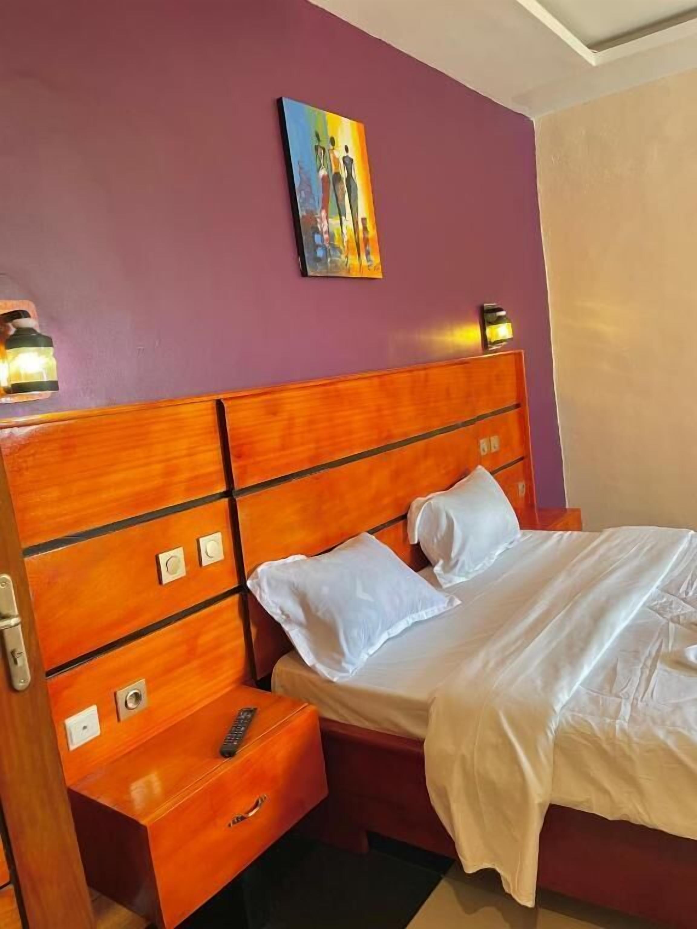 A hotel room with a wooden headboard, a bed with white linens, a nightstand with a remote control, and a painting on the wall.