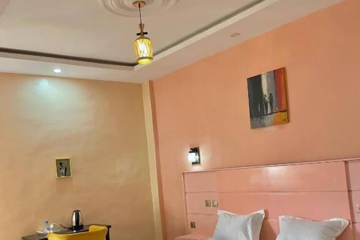 A bedroom with a pink bed, white bedding, a small table with a yellow chair, and a painting on the wall.