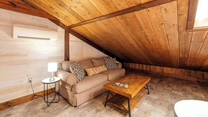 A cozy attic room with a wooden ceiling, a beige sofa with patterned cushions, a small side table with a lamp, and a wooden coffee table. - Spacious Barn Apt w/ King Bed (Asheville)