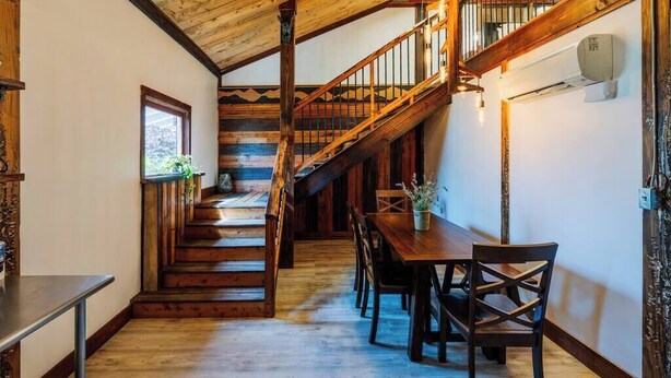 A wooden staircase leading to a second floor, a dining area with a wooden table and chairs, and a window with a view of greenery. - Spacious Barn Apt w/ King Bed (Asheville)