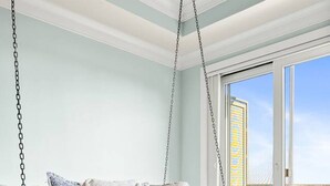 Ein Zimmer mit Hängesessel, einer Couch mit Kissen, einem großen Fenster und Blick ins Freie.