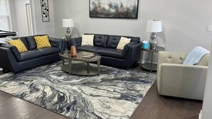 Ein Wohnzimmer mit einem dunkelbraunen Ledersofa, einem runden Couchtisch und einem Teppich mit Marmor-Muster. Zwei Beistelltische mit Lampen, ein beiges Sessel und ein Gemälde an der Wand sind ebenfalls zu sehen.