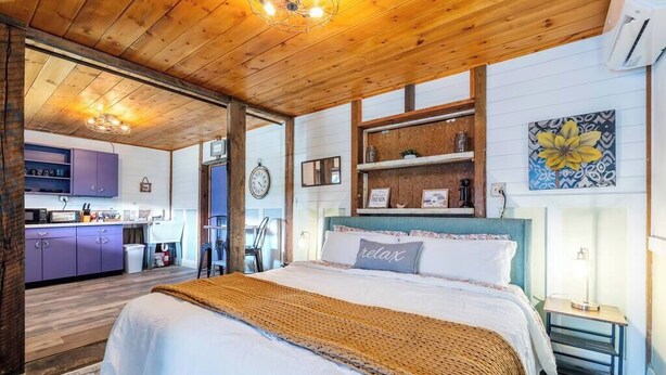 Ein gemütliches Schlafzimmer mit Holzboden, einem Bett mit beiger Decke, einem Nachttisch und einem Wandregal mit Dekoration.