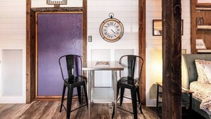 Ein Zimmer mit Holzbalkendecke, einer violetten Tür, einer runden Wanduhr, zwei schwarzen Stühlen, einem kleinen Tisch und einer Couch mit floralen Kissen.