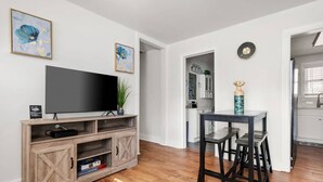 Ett modernt vardagsrum med en platt-TV på ett trästativ, en matplats med bord och stolar samt ett kök i bakgrunden.