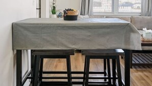 En matplats med ett bord täckt av en grå duk, trästolar, en väggklocka och ett fönster med gardiner.